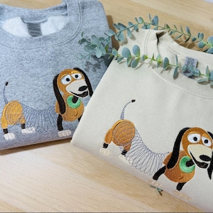 K&ouml;nnte beinhalten: Ein graumeliertes Sweatshirt und ein cremefarbenes Sweatshirt, jeweils mit einer Stickerei eines braun-beigen Dackel-Spielzeugs mit spiralf&ouml;rmigem K&ouml;rper, schwarzen Ohren und gr&uuml;nem Halsband, nebeneinander auf einer hellen Holzoberfl&auml;che platziert.