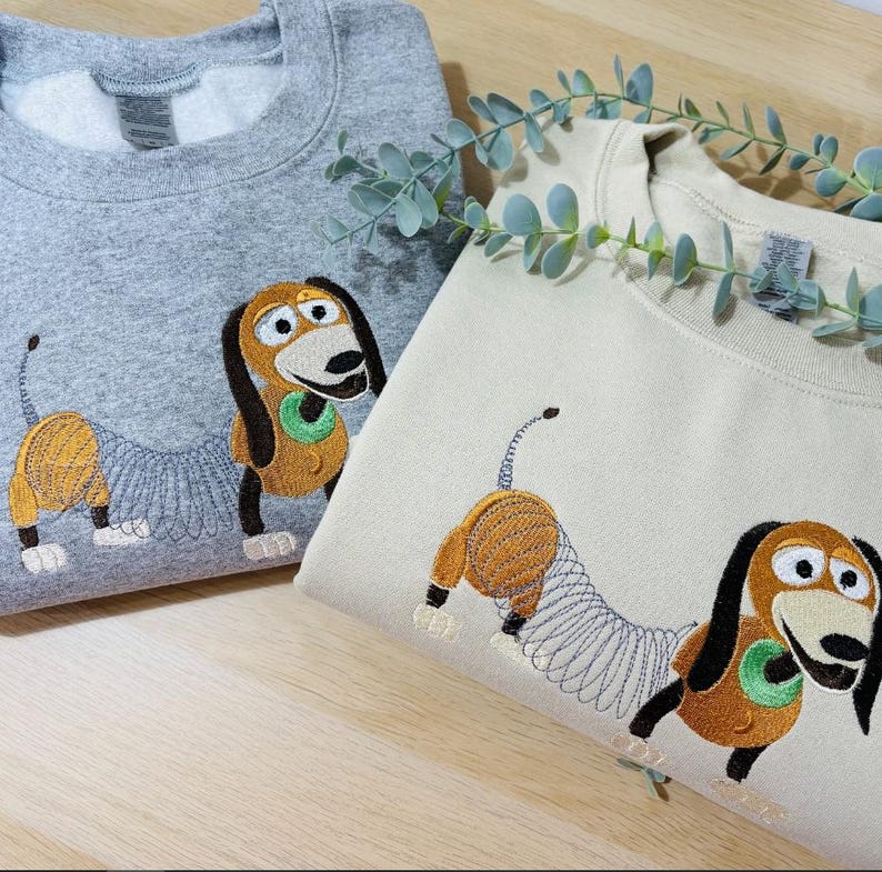 K&ouml;nnte beinhalten: Ein graumeliertes Sweatshirt und ein cremefarbenes Sweatshirt, jeweils mit einer Stickerei eines braun-beigen Dackel-Spielzeugs mit spiralf&ouml;rmigem K&ouml;rper, schwarzen Ohren und gr&uuml;nem Halsband, nebeneinander auf einer hellen Holzoberfl&auml;che platziert.
