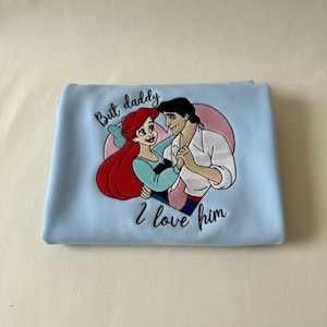 May include: Light blue blanket with an embroidered image of Ariel and Prince Eric from The Little Mermaid. The image is set against a pink heart, with the text "But daddy I love him" in black script.