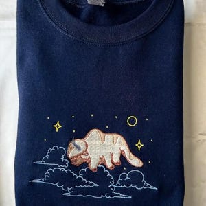 Può includere: Felpa blu navy con un design ricamato di una creatura simile a un bisonte, nuvole e stelle. Il design presenta tonalità beige, marrone e bianco, con stelle gialle e un cerchio giallo. La felpa è di colore blu navy.