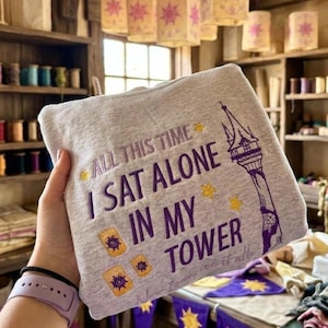Pode incluir: Um moletom cinza claro com o texto roxo "ALL THIS TIME I SAT ALONE IN MY TOWER". O design inclui uma ilustração de torre e símbolos de sol. O moletom é segurado em uma mão.