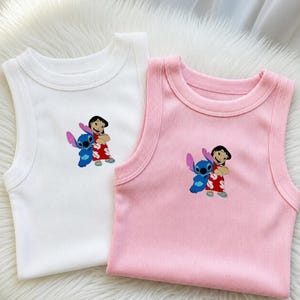 May include: Two ribbed tank tops, one white and one pink, featuring an embroidered image of Lilo and Stitch. The image shows Lilo in a red dress and Stitch in blue. The tops have a round neckline.