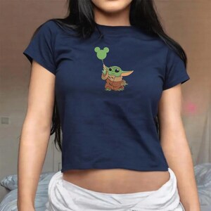 May include: A navy blue short-sleeved t-shirt featuring a graphic print of a small green creature with large ears wearing a brown robe. The creature is holding a green balloon shaped like a mouse head with circular ears and a striped pattern.