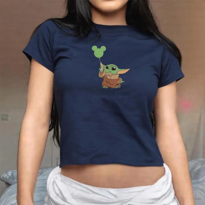 May include: A navy blue short-sleeved t-shirt featuring a graphic print of a small green creature with large ears wearing a brown robe. The creature is holding a green balloon shaped like a mouse head with circular ears and a striped pattern.