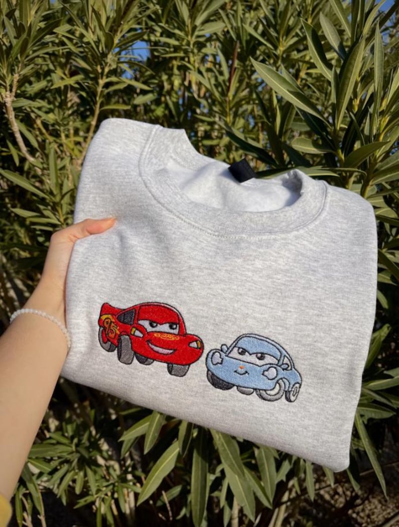 Puede incluir: Una sudadera gris claro con im&aacute;genes bordadas de dos coches de dibujos animados: un coche rojo con el n&uacute;mero 95 y un coche azul. La sudadera est&aacute; hecha de un material suave y tiene cuello redondo.