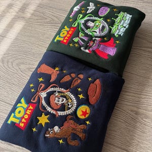 May include: Two embroidered sweatshirts featuring characters from the animated film Toy Story. One is dark green with Buzz Lightyear and other characters. The other is navy blue with Woody and other characters. Both have the Toy Story logo.