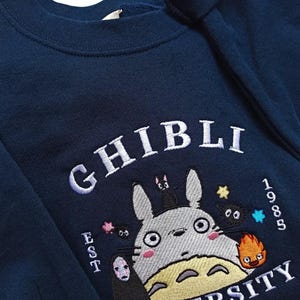 Pu&ograve; includere: Felpa blu navy con ricamo "Ghibli University". Presenta un personaggio dei cartoni animati con altre figure animate, il testo "EST 1985" e la parola "Ghibli" in bianco.