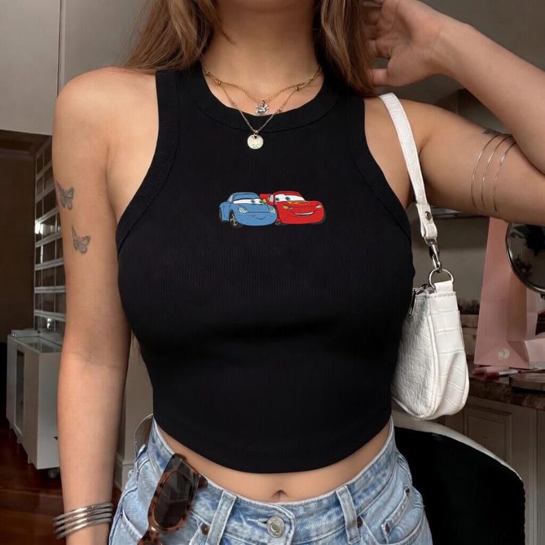 May include: Black ribbed tank top featuring an embroidered design of two cartoon cars, one blue and one red. The top has a round neckline and a fitted silhouette. The Cars-themed top is a casual and stylish piece.