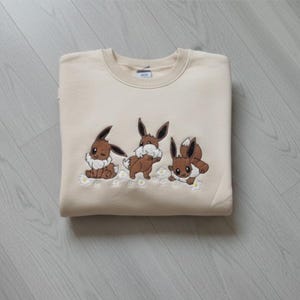 Op de afbeelding: Een beige sweatshirt met een geborduurd ontwerp van drie bruine stripfiguren met grote oren en pluizige staarten. De figuren zijn geplaatst op een rij kleine witte bloemen. De sweatshirt is netjes opgevouwen.