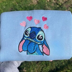 Puede incluir: Sudadera azul claro con un bordado de Stitch, el personaje de Lilo & Stitch. Stitch es azul con orejas rosas y est&aacute; rodeado de corazones rosas. La sudadera es de material suave.