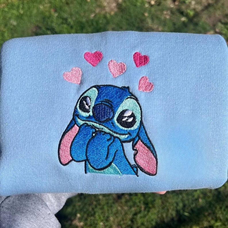Puede incluir: Sudadera azul claro con un bordado de Stitch, el personaje de Lilo & Stitch. Stitch es azul con orejas rosas y est&aacute; rodeado de corazones rosas. La sudadera es de material suave.