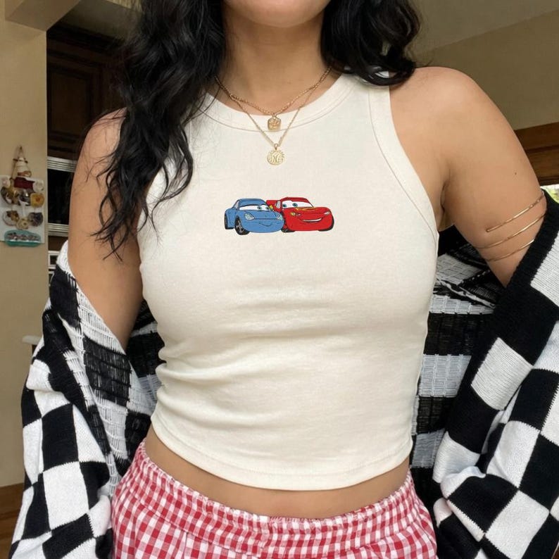 May include: Cream-colored tank top featuring a graphic of two cartoon cars, one blue and one red. The top is a sleeveless, ribbed style. The person is wearing a black and white checkered cardigan and red and white checkered shorts.