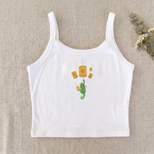 May include: White tank top with a colorful graphic featuring floating lanterns and a green seahorse-like creature. The design includes yellow, orange, and green elements. The top has a classic, sleeveless design.