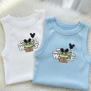 May include: Two baby tank tops, one white and one light blue, featuring an embroidered design. The design includes a green alien character wearing a Mickey Mouse hat and holding a black balloon, with the text "W" and "WARS".