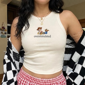 K&ouml;nnte beinhalten: Cremefarbenes Tanktop mit einem Cartoon-Bild von Woody aus Toy Story und dem Wort "overstimulated". Das Top ist &auml;rmellos und hat eine gerippte Textur.