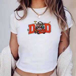 K&ouml;nnte beinhalten: Wei&szlig;es T-Shirt mit einem Aufdruck des Wortes "DAD" in roten Blockbuchstaben und Zeichentrickautos. Das Design enth&auml;lt ein Route-66-Emblem und eine Troph&auml;e. Die Autos sind rot, braun und blau.