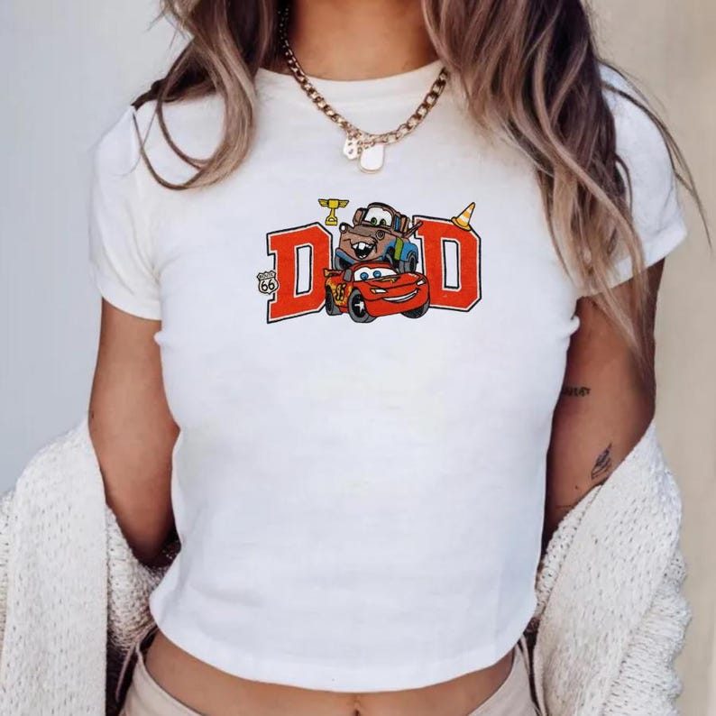 K&ouml;nnte beinhalten: Wei&szlig;es T-Shirt mit einem Aufdruck des Wortes "DAD" in roten Blockbuchstaben und Zeichentrickautos. Das Design enth&auml;lt ein Route-66-Emblem und eine Troph&auml;e. Die Autos sind rot, braun und blau.