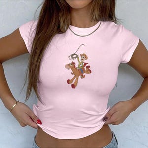 May include: A light pink t-shirt featuring a cartoon graphic of a cowboy riding a horse. The cowboy is wearing a hat and holding a lasso. The horse is brown with red hooves. The shirt has short sleeves.