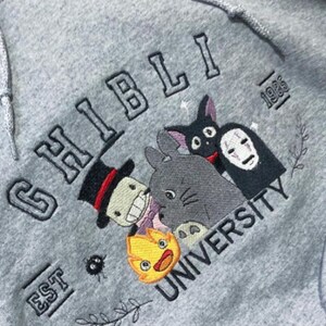 Puede incluir: Sudadera con capucha gris con dise&ntilde;o bordado "Ghibli University". Presenta personajes de las pel&iacute;culas de Studio Ghibli, como Totoro, Sin Cara y Jiji. Tambi&eacute;n incluye el texto "EST 1985".