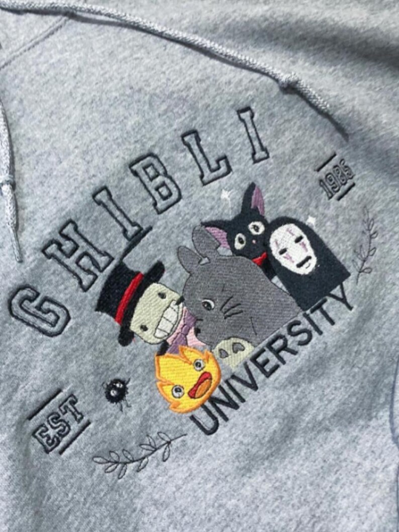 Puede incluir: Sudadera con capucha gris con dise&ntilde;o bordado "Ghibli University". Presenta personajes de las pel&iacute;culas de Studio Ghibli, como Totoro, Sin Cara y Jiji. Tambi&eacute;n incluye el texto "EST 1985".