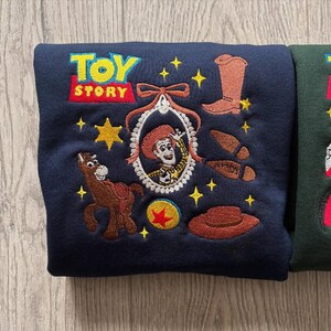 May include: Navy blue sweatshirt with embroidered Toy Story characters. Features the Toy Story logo, Jessie, a horse, a cowboy boot, a hat, and other elements from the movie. The sweatshirt is a comfortable, casual garment.