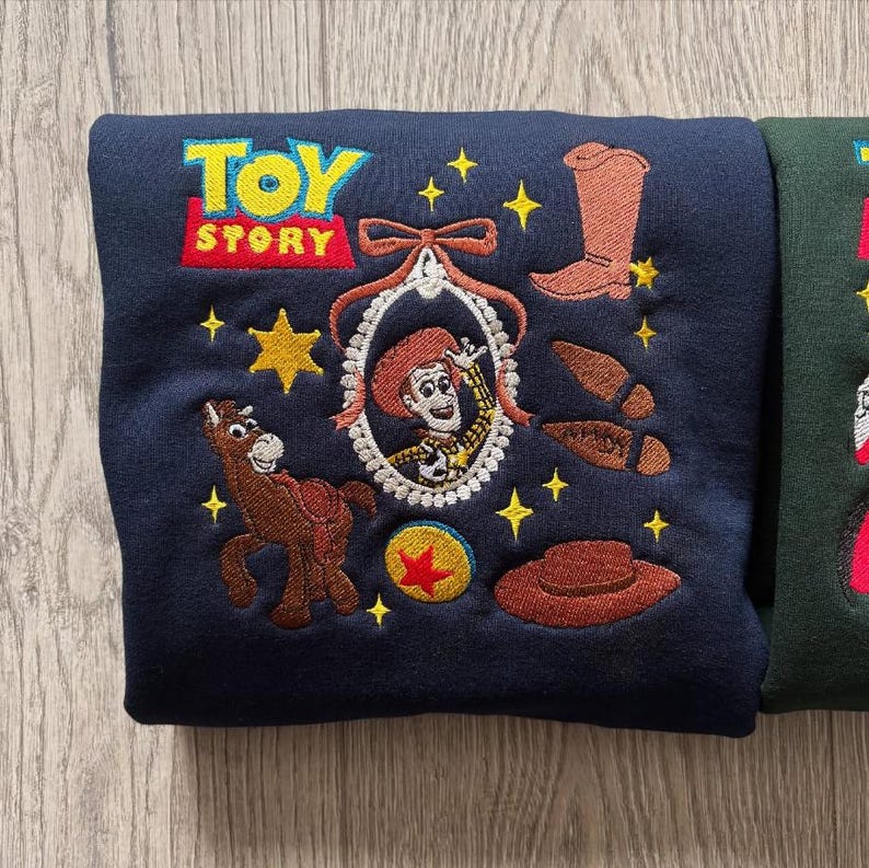May include: Navy blue sweatshirt with embroidered Toy Story characters. Features the Toy Story logo, Jessie, a horse, a cowboy boot, a hat, and other elements from the movie. The sweatshirt is a comfortable, casual garment.