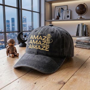 May include: A gray baseball cap with the word "AMAZE" in gold lettering. The cap has a curved brim and a distressed look. The hat is sitting on a wooden surface.