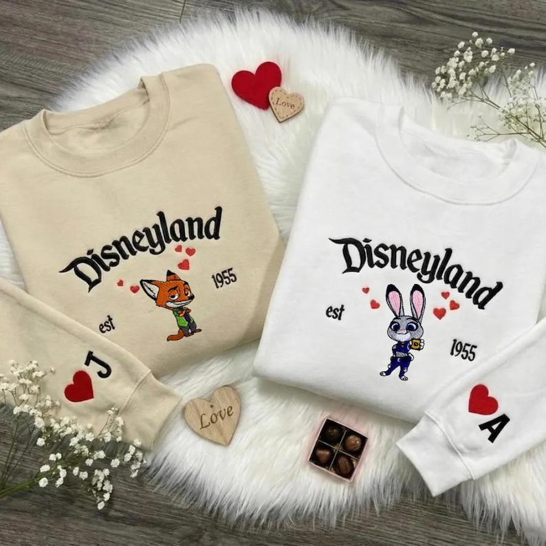 Op de afbeelding: Twee crewneck sweatshirts, &eacute;&eacute;n beige en &eacute;&eacute;n wit, met "Disneyland" geborduurd in zwart script. Elk heeft een Disney-personage en het jaar "1955". Mouwen tonen speelkaartsymbolen. Een houten decoratie in de vorm van een hart is ook zichtbaar.