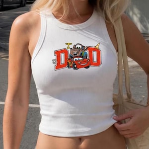 K&ouml;nnte beinhalten: Wei&szlig;es geripptes Tanktop mit einer farbenfrohen Grafik von Zeichentrickautos und dem Wort "DAD" in roten Blockbuchstaben. Die Grafik enth&auml;lt ein Route 66-Schild und eine Troph&auml;e.