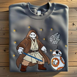 May include: Gray sweatshirt featuring embroidered Star Wars characters. The design includes a figure with a lightsaber, a BB-8 droid, and the Millennium Falcon, with yellow star accents.