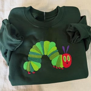 Peut inclure: Sweat-shirt vert fonc&eacute; avec un motif de chenille brod&eacute;e. La chenille a une t&ecirc;te rouge, des yeux noirs, des antennes violettes et un corps segment&eacute; vert et jaune. Le sweat-shirt est en mati&egrave;re souple.