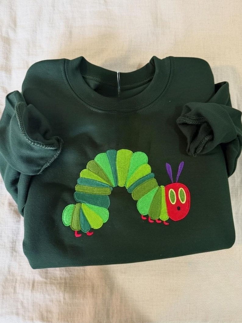 Peut inclure: Sweat-shirt vert fonc&eacute; avec un motif de chenille brod&eacute;e. La chenille a une t&ecirc;te rouge, des yeux noirs, des antennes violettes et un corps segment&eacute; vert et jaune. Le sweat-shirt est en mati&egrave;re souple.