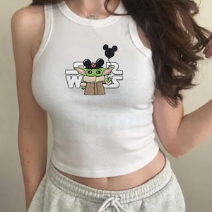 May include: White tank top with a graphic of Grogu from Star Wars wearing Mickey Mouse ears and holding a Mickey Mouse balloon. The Star Wars logo is also featured. The top is sleeveless and has a round neckline.