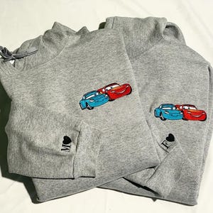 May include: Two grey sweatshirts featuring embroidered cartoon car designs. One has the letter "M" and a heart on the sleeve, the other has the letter "F" and a heart. The cars are blue and red.