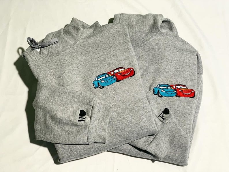 May include: Two grey sweatshirts featuring embroidered cartoon car designs. One has the letter "M" and a heart on the sleeve, the other has the letter "F" and a heart. The cars are blue and red.