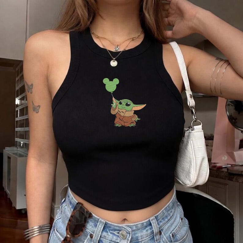 May include: A black ribbed tank top featuring a graphic print of a small green creature with large ears wearing a brown robe and holding a green balloon shaped like a mouse head. The top has a high neckline and a fitted, sleeveless silhouette.