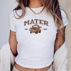 May include: A white short-sleeved t-shirt featuring a graphic of a brown tow truck character. The text MATER is printed in a brown collegiate font above the truck, with EST. 1977 printed on either side of the truck graphic in a smaller brown font.