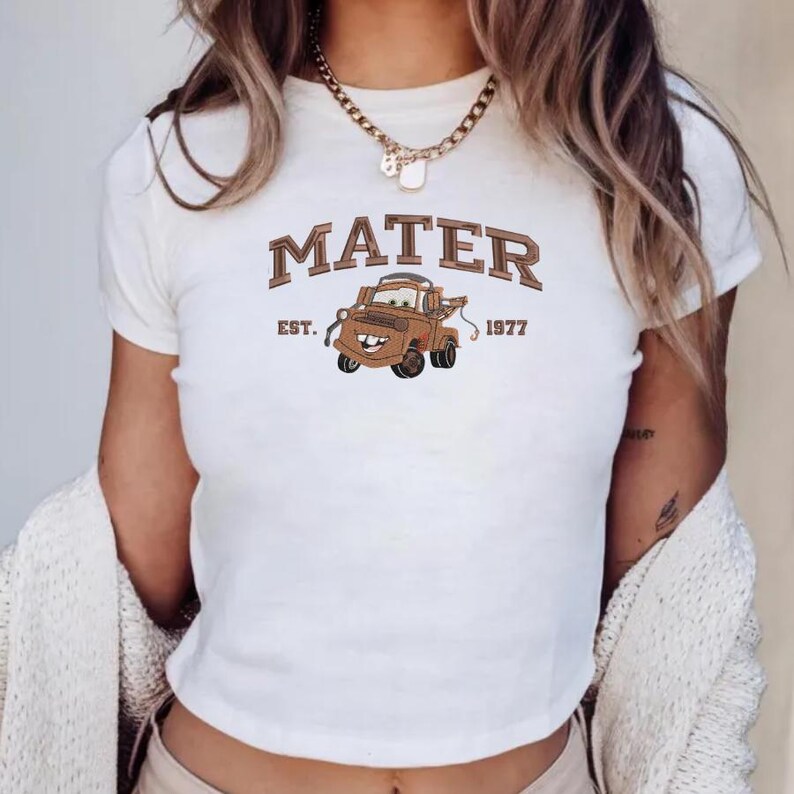 May include: A white short-sleeved t-shirt featuring a graphic of a brown tow truck character. The text MATER is printed in a brown collegiate font above the truck, with EST. 1977 printed on either side of the truck graphic in a smaller brown font.