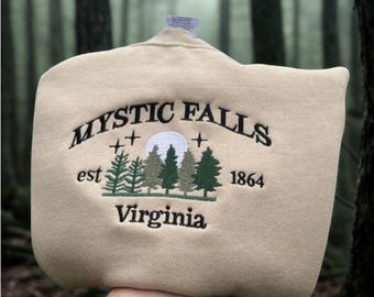 May include: A tan sweatshirt with the embroidered text "MYSTIC FALLS Virginia 1864" above a forest scene. The design includes a moon and trees in shades of green. The sweatshirt is held up against a blurred forest background.