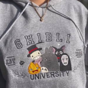 Puede incluir: Sudadera gris con dise&ntilde;o bordado "Ghibli University". Presenta varios personajes de dibujos animados en un dise&ntilde;o colorido. La sudadera tiene cord&oacute;n y bolsillo delantero. El texto "EST 1985" tambi&eacute;n est&aacute; bordado.