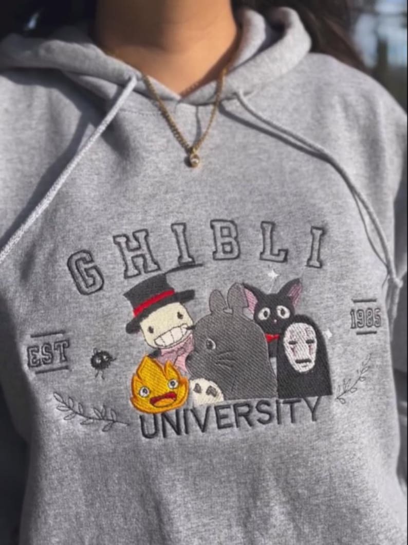 Puede incluir: Sudadera gris con dise&ntilde;o bordado "Ghibli University". Presenta varios personajes de dibujos animados en un dise&ntilde;o colorido. La sudadera tiene cord&oacute;n y bolsillo delantero. El texto "EST 1985" tambi&eacute;n est&aacute; bordado.