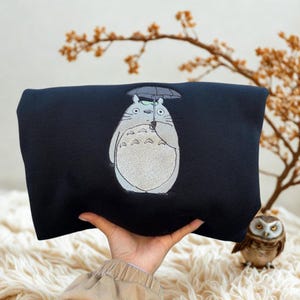 May include: A navy blue sweatshirt featuring an embroidered image of Totoro holding an umbrella. The design is in shades of gray, white, and black. The sweatshirt is held up by a person's hand.