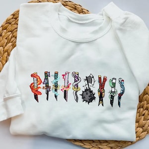 Puede incluir: Sudadera blanca de cuello redondo con cuchillos bordados de colores. Los cuchillos tienen varios diseños, incluyendo patrones florales y geométricos. La sudadera se muestra sobre una superficie tejida de color marrón claro.