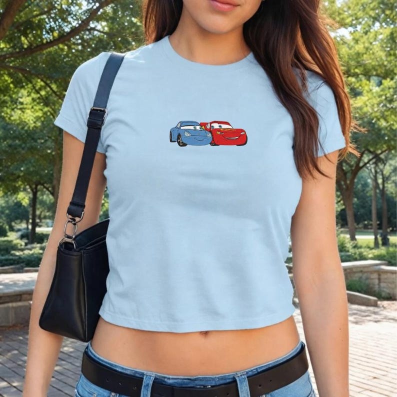 May include: Light blue crop top featuring a cartoon graphic of two race cars, one blue and one red. The cars are smiling and appear to be embracing. The shirt is made of a soft material and has short sleeves.