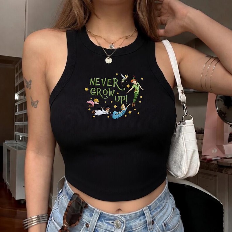 Peut inclure: Un d&eacute;bardeur noir c&ocirc;tel&eacute; avec une impression graphique des personnages de Peter Pan et le texte NEVER GROW UP! entour&eacute; de petites &eacute;toiles jaunes. Le haut sans manches pr&eacute;sente une encolure ronde et un ourlet court, avec un sac blanc.