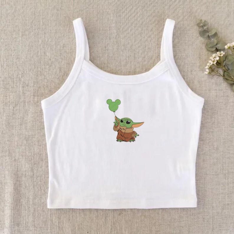 May include: A white sleeveless crop top featuring a central graphic of a small green creature with large ears wearing a brown robe and holding a light green balloon shaped like a mouse head. The garment has a scoop neckline and wide shoulder straps.