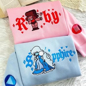 Op de afbeelding: Twee sweatshirts, één roze en één lichtblauw, met geborduurde cartoonfiguren en tekst. Het roze sweatshirt zegt "Ruby" met een cartoonfiguur. Het lichtblauwe sweatshirt zegt "Sapphire" met een cartoonfiguur.