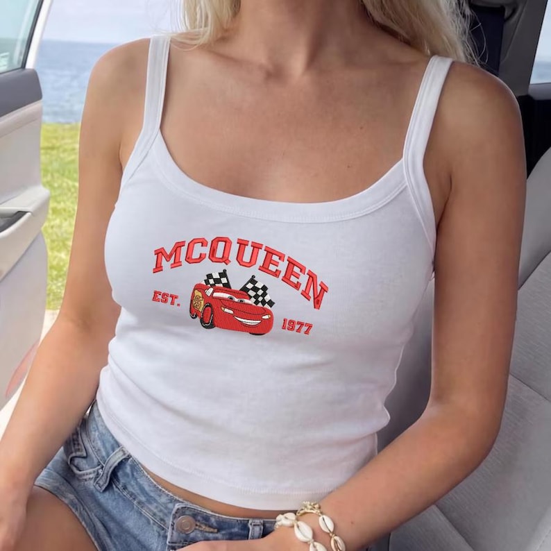 Puede incluir: Camiseta sin mangas blanca con la palabra "MCQUEEN" en rojo, junto con una imagen de dibujos animados de un coche de carreras rojo y banderas a cuadros. El texto "EST. 1977" tambi&eacute;n est&aacute; impreso en la camiseta. La camiseta sin mangas tiene cuello redondo y tirantes finos.