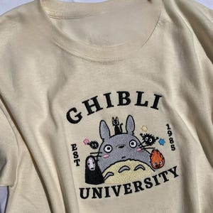 Pu&ograve; includere: T-shirt color crema con un design ricamato. Il design presenta le parole "GHIBLI UNIVERSITY" e l'anno "1985". Il design include un personaggio dei cartoni animati grigio, una piccola figura nera e altri personaggi animati colorati.