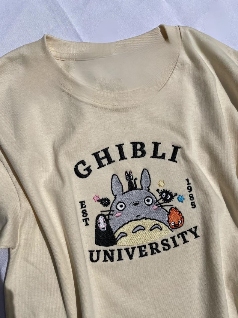 Pu&ograve; includere: T-shirt color crema con un design ricamato. Il design presenta le parole "GHIBLI UNIVERSITY" e l'anno "1985". Il design include un personaggio dei cartoni animati grigio, una piccola figura nera e altri personaggi animati colorati.
